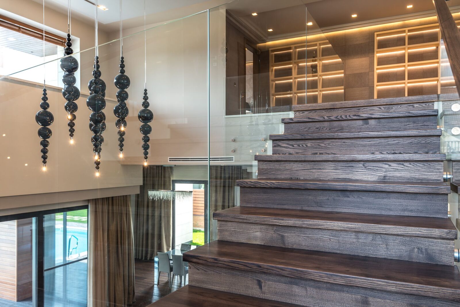 Dark timber stair rising beside a frameless glass balustrade under pendant lights